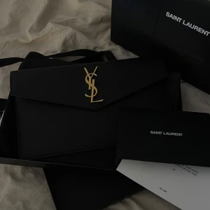 Svart kuvertväska från Saint Laurent - Säljer en svart kuvertväska från Saint Laurent med ikoniskt YSL-emblem i guld på framsidan. Väskan har en stilren design med axelrem och är tillverkad i skinn. Väskan är i toppskick och utan skrapor! Kommer med kvitto och dustbag.