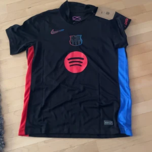 FC Barcelona herrtröja Lamine Yamal 19 - Säljer en svart FC Barcelona fotbollströja med röda och blå detaljer från Nike. Tröjan har korta ärmar, Spotify-logga på framsidan och Lamine Yamal 19 på ryggen. Materialet är lätt och andas, perfekt för match eller träning.