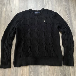 Trändig Svart kabelstickad tröja från Polo Ralph Lauren - Kabelstickad svart tröja från Polo Ralph Lauren med rund halsringning och broderad logga på bröstet. Klassisk modell med ribbade muddar vid ärmslut och nederkant. Perfekt för lager-på-lager under kyliga dagar.