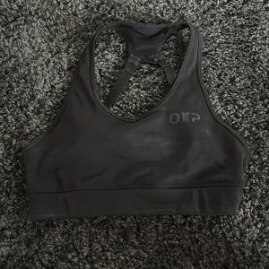 Svart sport-bh från OTP - Säljer en svart sport-bh från OTP med brottarrygg och justerbara axelband. Perfekt för träning och ger bra stöd. Snygg och enkel design med diskret logga framtill.