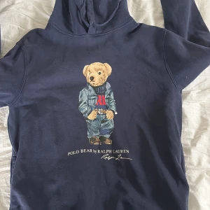 Mörkblå hoodie med Polo Bear från Ralph Lauren - Säljer en mörkblå hoodie från Ralph Lauren med den ikoniska Polo Bear-trycket på framsidan. Tröjan har huva och långa ärmar, perfekt för en avslappnad och cool stil. Klassisk och tidlös design med mjukt material.