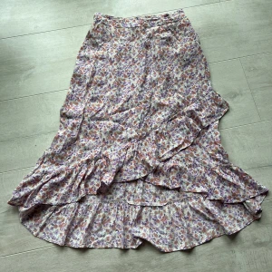 Blommig volangkjol från Pieces - Säljer en midi kjol från Pieces med blommigt mönster i lila, rosa och orange toner. Kjolen har volangdetaljer och är tillverkad i ett lätt och luftigt material, perfekt för varmare dagar.