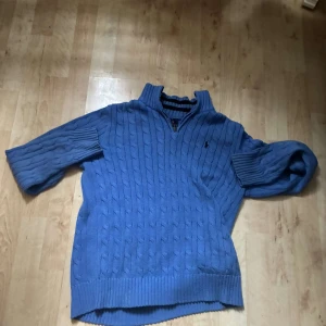 Blå stickad tröja med dragkedja från Polo Ralph Lauren - Säljer en blå stickad tröja från Polo Ralph Lauren med halv dragkedja och kabelstickat mönster. Tröjan har hög krage och långa ärmar, perfekt för kyligare dagar. Klassisk och stilren design med broderad logga på bröstet.