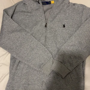 Grå Polo Ralph Lauren zip  - Säljer en grå zip-hoodie från Polo Ralph Lauren med klassisk broderad logga på bröstet. Jackan har huva med dragsko, långa ärmar och ribbade muddar. Perfekt för en avslappnad och stilren look.