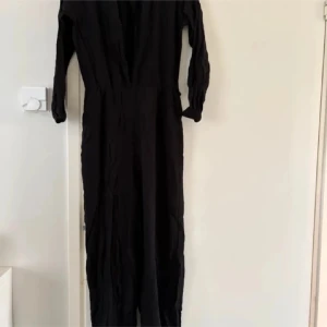 Svart jumpsuit med v-ringning från H&M - Svart jumpsuit från H&M med djup v-ringning och långa ärmar. Den har en enkel och stilren design med markerad midja och lätt rynkade detaljer. Perfekt för dig som gillar minimalistisk stil. Strl S
