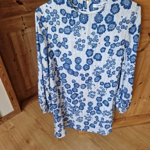 Blommig klänning från Samsøe Samsøe - Superfin klänning från Samsøe Samsøe med blått blommönster på vit botten. Klänningen har lång ärm, rund halsringning och dragkedja bak. Tillverkad i viskos och polyester för en lätt och skön känsla.