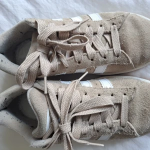 Beige Adidas Campus sneakers - Säljer ett par beige Adidas Campus sneakers med vita ränder och gummisula. Skorna har klassisk snörning och är i mocka med Adidas-logga bak. Perfekta för dig som gillar stilrena och tidlösa sneakers. Använda fåtal gånger, dock saknas innersula då jag haft egen och hittar inte originalet,men funkar att lägga i vilken sula man vill. Jätte fina.