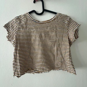 Randig croptop från Pull&Bear - Säljer en croppad t-shirt från Pull&Bear i vitt med tunna blå och senapsgula ränder. T-shirten har korta ärmar och en rund halsringning. Perfekt till sommaren och enkel att matcha med jeans eller kjol. Jättefint skick, knappt använd. Passar XS/S.