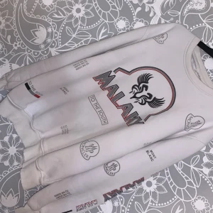 Vit sweatshirt med tryck från Malay - Vit sweatshirt med svart och rött tryck framtill och på ärmarna. Stort grafiskt motiv och texten 'MALAY' på bröstet. Rund halsringning och ribbade muddar vid ärmslut och nederkant. Perfekt för en avslappnad stil.