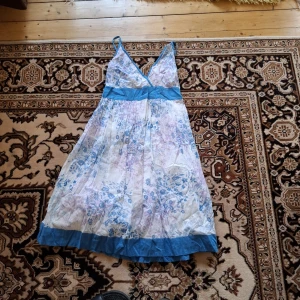 Blommig sommarklänning med blå detaljer - Söt och luftig klänning med blommigt mönster i blått och lila på vit botten. Klänningen har v-ringning, breda axelband och markerad midja med blå band. Den är ärmlös och har en lätt utställd kjol med blå kant nertill. Perfekt för varma dagar.