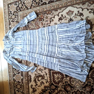 Randig långklänning i bomull - Säljer en luftig långklänning med blå och vita ränder. Klänningen har lång ärm och volangdetaljer nertill. Perfekt för varma dagar och har en avslappnad passform. Materialet känns mjukt och svalt mot huden.