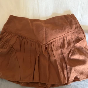 Rostbrun kortkjol med volang - Säljer en rostbrun kortkjol med volangdetaljer och bred linning med shorts under. Kjolen har en avslappnad passform och är tillverkad i ett mjukt, lätt material som ger ett snyggt fall. Aldrig använd