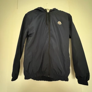 Blå vindjacka från Moncler - Säljer en mörkblå vindjacka från Moncler med huva och dragkedja framtill. Jackan har Moncler-logga på bröstet och elastiska muddar vid ärmslut och nederkant. Perfekt för blåsiga dagar och enkel att matcha med olika outfits.
