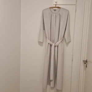 Ljusgrå/ljusblå långklänning med bälte från COS - Säljer en stilren ljusgrå/ljusblå långklänning från COS med lång ärm och knytbälte i midjan. Klänningen har rund halsringning och ett minimalistiskt snitt som ger en modern look.