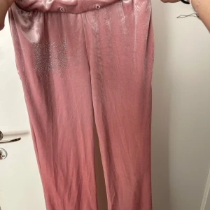 Rosa mjukisbyxor med strassdetalj - Säljer ett par glansiga rosa mjukisbyxor med resår i midjan och en snygg strassdetalj på ena benet. Perfekta för en chill och trendig look.