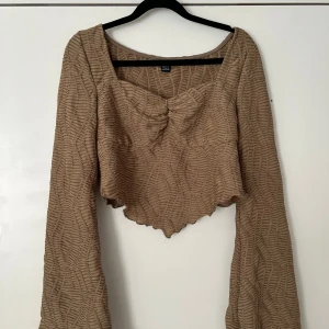 Beige croppad topp med lång ärm - Trendig beige croppad topp med lång ärm och vågig kant nertill. Toppen har en rynkad detalj framtill och är tillverkad i ett strukturerat, stretchigt material. Perfekt för att skapa en cool och avslappnad look.