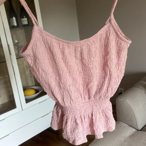 Ljusrosa smockad linnetopp - Säljer en ljusrosa linnetopp med smockad struktur och tunna axelband. Toppen har en volang nertill som ger en söt och somrig känsla. Perfekt att matcha med jeans eller kjol för en avslappnad look.