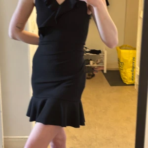 Svart volangklänning från ELA - Säljer en svart kortklänning från ELA med volangdetaljer vid axlar och fåll. Klänningen har en tight passform och är ärmlös, perfekt för dig som vill ha en stilren och elegant look. Passar även i storlek S