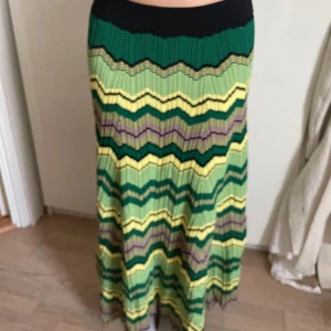Mönstrad långkjol från Sandro Paris - Säljer en långkjol från Sandro Paris med vågigt mönster i grönt, gult, svart och lila. Kjolen har elastisk midja och är stickad i ett mjukt material. Perfekt för dig som gillar färg och retrovibbar!