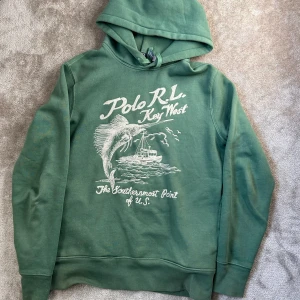 Grön hoodie från Polo Ralph Lauren - Säljer en grön hoodie från Polo Ralph Lauren med tryck framtill med texten 'Polo RL Key West'. Tröjan har huva med dragsko och ribbade muddar vid ärmslut och nederkant. Tröjan är mycket sällsynt och nästintill oanvänd.