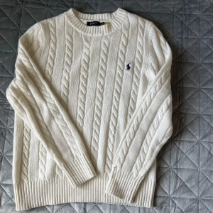 Vit kabelstickad tröja från Polo Ralph Lauren - Kabelstickad vit tröja från Polo Ralph Lauren med rund halsringning och broderad marinblå logga på bröstet. Klassisk modell med långa ärmar och ribbade muddar. Perfekt för lager-på-lager och stilrena outfits.