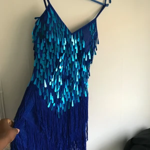 Blå paljettklänning med fransar - Säljer en blå klänning med smala axelband, dekorerad med glittriga paljetter och långa fransar framtill. Perfekt för dig som vill sticka ut på dansgolvet! Klänningen har en figurnära passform och är tillverkad i ett glansigt material.