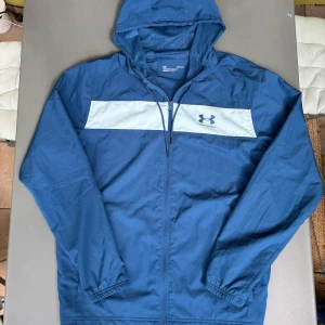 Blå vindjacka med huva från Under Armour - Säljer en blå vindjacka från Under Armour med vit panel och logga på bröstet. jackan är ny skick. Inga skador. Allt funkade bra jättesnygg passform perfekt att ha till sommaren