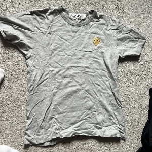 Grå t-shirt från Comme des Garçons Play - Säljer en grå t-shirt från Comme des Garçons Play med det ikoniska gula hjärtat broderat på bröstet. T-shirten har rund hals och korta ärmar. Perfekt för en stilren och trendig look.