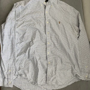 Rutig skjorta från Ralph Lauren - Klassisk rutig skjorta från Ralph Lauren i vitt, blått och svart. Skjortan har lång ärm, knappar framtill och broderad logga på bröstet. Perfekt för dig som gillar tidlös stil och vill ha något från ett ikoniskt märke.