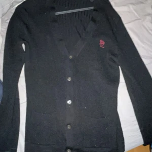 Svart kofta från Alexander McQueen - Säljer en svart stickad kofta från Alexander McQueen med knappar framtill och två fickor. Koftan har en broderad röd logga på bröstet och v-ringad hals. Perfekt för dig som gillar stilrena och exklusiva plagg.