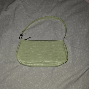 Ljusgrön axelväska med krokodilpräglat mönster - Säljer en ljusgrön axelväska i syntetmaterial med krokodilpräglad yta. Väskan har ett kort axelband och stängs med dragkedja upptill. Perfekt för att bära det viktigaste och ger en trendig touch till din outfit.