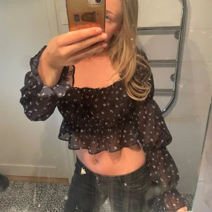 Svart blommig croppad blus med volang - Säljer en svart croppad blus med små vita blommor. Blusen har långa, transparenta ärmar med resår vid handlederna och volangdetaljer både vid ärmslut och nederkant. Perfekt för sommaren. Väldigt stretchig så passar d flesta!