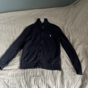Mörkblå stickad kofta från Polo Ralph Lauren - Säljer en mörkblå stickad kofta från Polo Ralph Lauren med dragkedja framtill och ribbade muddar.Passar mellan 149-159 cm,org pris 1800kr mitt pris 400kr, priset är ej hugget i sten