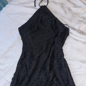 Svart spetsklänning med smala axelband runt halsen  - Säljer en svart klänning med smala axelband runt halsen. Klänningen har vacker spets med blommigt mönster och en dragkedja i sidan. Den har en figurnära passform som framhäver siluetten och blir rak i slutet har underkjol till precis över knäna 