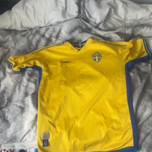 Svenska landslagets fotbollströja från Umbro - Gul kortärmad fotbollströja från Umbro med blå detaljer och svenska landslagets emblem på bröstet. Klassisk V-ringning med blå och gul krage, samt blå sidor. Perfekt för dig som vill visa ditt stöd för Sverige!