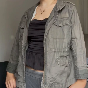 Grå utilityjacka med fickor - Säljer en grå utilityjacka med flera stora fickor framtill och klassisk krage. Jackan har lång ärm och dragkedja. Perfekt för en avslappnad och cool stil.