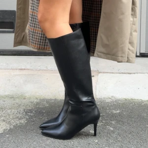 Svarta knähöga stövlar med klack från Nelly - Säljer ett par svarta knähöga stövlar från Nelly med spetsig tå och hög smal klack som för tillfället är slutsålda och inte i lager! Skorna har en stilren design som passar perfekt till årets alla tillfällen och outfits.