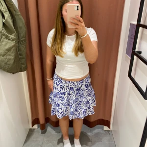 Vit och blå mönstrad kjol från Gina Tricot - Säljer en superfin vit kjol med blått blommigt mönster från Gina Tricot. Kjolen är kort, har volanger och resår i midjan för extra komfort. Perfekt för sommarens alla tillfällen! 💙