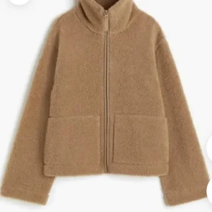 Beige teddyjacka med dragkedja - Mysig beige teddyjacka med hög krage och dragkedja framtill. Jackan har två stora fickor och är perfekt för kyliga dagar. Den har en rak passform och är helfärgad utan mönster.