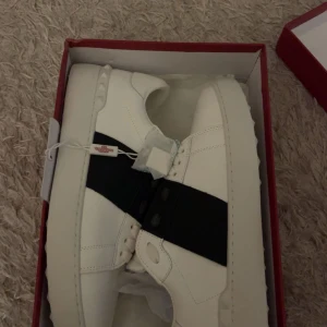 Valentino Garavani vita sneakers med svart band - Säljer ett par vita Valentino Garavani sneakers med en bred svart rem över mitten. Skorna har rund tå, platt sula och är tillverkade i skinn. Klassisk och stilren design med diskret logga på sidan.