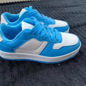 Blå och vita sneakers med snörning - Säljer ett par fräscha sneakers i blått och vitt med klassisk design. Skorna har rund tå, platt sula och är tillverkade i syntetmaterial. Perfekta för dig som gillar sportig stil och vill ha bekväma skor till vardagen.