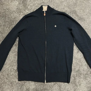 Mörkblå kofta från Morris - Säljer en stilren mörkblå kofta från Morris i 100% merinoull. Koftan har hel dragkedja framtill, ribbade muddar och en liten broderad logga på bröstet. Perfekt för lager-på-lager och snygg till både jeans och chinos.