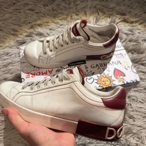 Dolce & Gabbana skor - Säljer ett par snygga vita sneakers från Dolce & Gabbana med vinröda detaljer på häl och plös. Skorna har snörning, rund tå och platt sula. Allt som syns på bilderna kommer med. De står Dolce Gabbana på baksidan av skon. Priset är lite lägre eftersom de finns en pytte liten skada på ena skon,som man ser på sista bilden. 7/10 skick. Pris kan diskuteras.Bara att höra av sig vid minsta fundering!