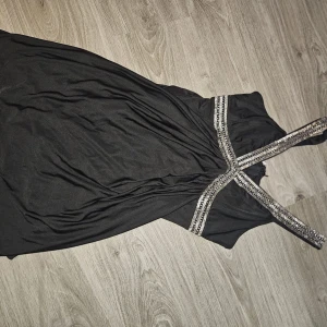 Svart långklänning med glittriga axelband - Säljer en svart långklänning med snygga glittriga axelband som korsas i ryggen. Klänningen har en elegant och stilren design, perfekt för speciella tillfällen. Materialet känns mjukt och följsamt.