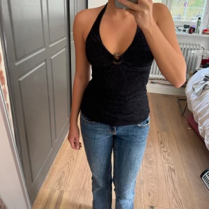 Svart halterneck linnetopp från Ellos - Snygg svart halterneck linnetopp från Ellos med virkad spets och dekorativ ring framtill. Toppen knyts i nacken och har en figurnära passform. Perfekt till jeans eller kjol för en trendig look.