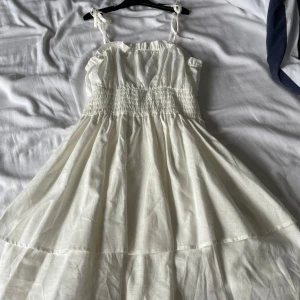 Vit sommarklänning med smock och volang - Säljer en söt vit klänning med smala axelband som knyts och smockad midja. Klänningen har volangdetaljer upptill. Perfekt för varma dagar och har en romantisk känsla.