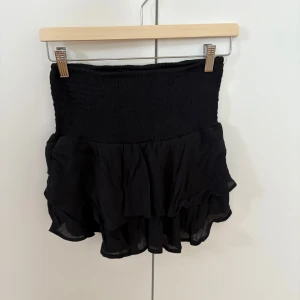 Svart volangkjol - Supersöt svart kortkjol med volanger och smock i midjan. Kjolen har en luftig och flowy känsla. Kan användas som både lågmidjad och högmidjad