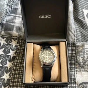 Seiko herrklocka med svart skinnarmband - Stilren Seiko-klocka med svart skinnarmband och silverfärgad boett. Urtavlan är grå med flera små urtavlor och tydliga markeringar. Klockan har klassisk design och levereras i originalask. Inga repor eller så