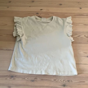 Beige ribbad topp med volangärmar från Zara - Säljer en beige ribbad topp från Zara i storlek 152 (11-12 år). Toppen har korta ärmar med volangdetaljer som ger en söt och trendig look. Perfekt för dig som gillar stilrena plagg med en twist!