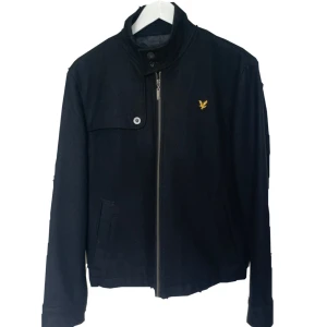 Svart jacka från Lyle & Scott - Snygg svart jacka från Lyle & Scott med gul logga på bröstet. Jackan har ståkrage, dragkedja framtill och en knappdetalj vid axeln. Perfekt för dig som vill ha en stilren look.